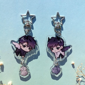 Puede incluir: Dos llaveros acrílicos con personajes de estilo anime con cabello oscuro y alas. Cada llavero tiene un cierre en forma de estrella y un pequeño dije con forma de fantasma. El fondo es azul claro.