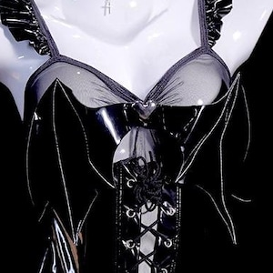 May include: Black lingerie top with bat wing accents. The top features a sheer mesh panel and a lace-up front. The straps have a ruffled trim. The bustier is displayed on a white mannequin against a black background.