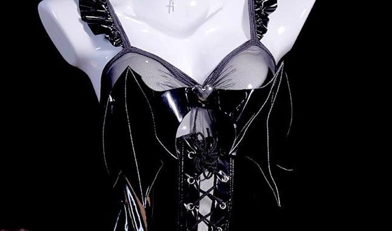 May include: Black lingerie top with bat wing accents. The top features a sheer mesh panel and a lace-up front. The straps have a ruffled trim. The bustier is displayed on a white mannequin against a black background.