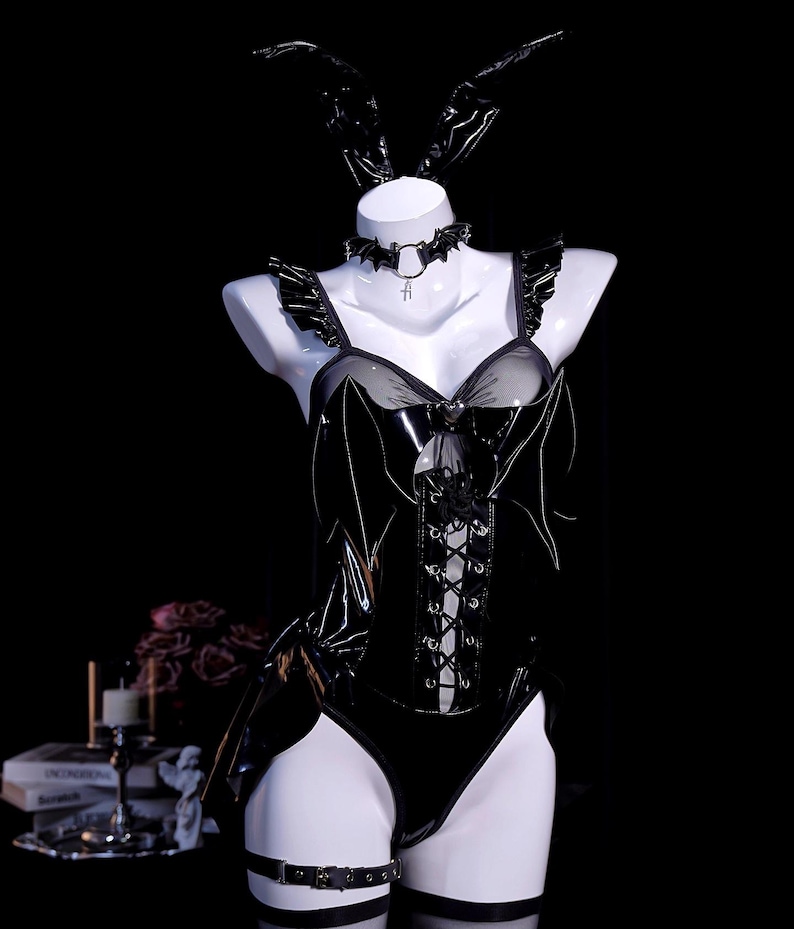 May include: A black, shiny bunny costume displayed on a mannequin. The costume includes a corset with lace-up detailing, a choker with a bat pendant, and a pair of bunny ears. A garter belt is visible on the thigh.