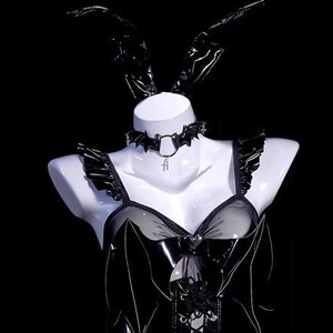 May include: Black lingerie set on a white mannequin. The set includes a bustier with a mesh panel, a bat-shaped choker, and matching bunny ears. The outfit has a gothic aesthetic.