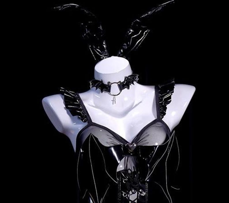 May include: Black lingerie set on a white mannequin. The set includes a bustier with a mesh panel, a bat-shaped choker, and matching bunny ears. The outfit has a gothic aesthetic.