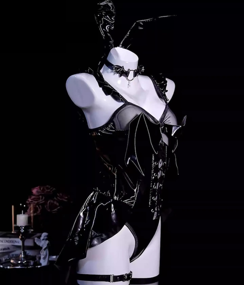 May include: A black, shiny bodysuit with a corset-style front, featuring a bat-wing design and a matching choker. The outfit includes a garter belt and bunny ears, displayed on a white mannequin against a dark background.