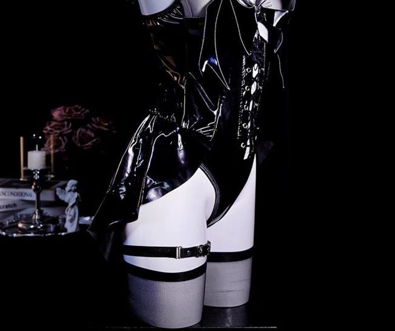 May include: Black patent leather bodysuit with lace-up detailing and a garter belt. The garment has a shiny, reflective surface and is displayed on a mannequin. The background is dark, emphasizing the product's sleek design.