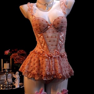 May include: A pink lingerie set on a mannequin. The set includes a corset with cherry print, a ruffled skirt, and matching garter bows. A pearl necklace adorns the mannequin's neck. The background is black.