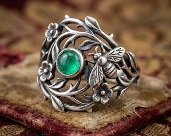 May include: A silver ring featuring an intricate openwork design of winding vines, leaves, and small flowers. A single round, polished green gemstone is set within the metalwork, accompanied by a detailed relief of a bee resting on the foliage.