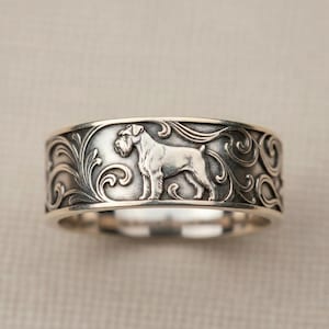 May include: A silver-toned metal ring featuring a detailed relief of a standing Schnauzer dog. The band is decorated with intricate, flowing scrollwork patterns that surround the dog figure, creating a textured and dimensional appearance.