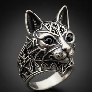 May include: A silver-colored ring featuring a detailed cat face design. The cat's face has black gemstone eyes and intricate patterns on the head and band. The ring has a gothic aesthetic.