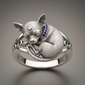 May include: A silver ring featuring a sleeping chihuahua with a blue collar and a diamond on its forehead. The ring band has emerald accents. The dog is curled up, with its eyes closed. A unique piece of jewelry.