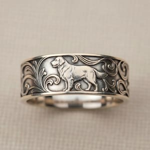 May include: Silver-toned ring featuring a detailed relief of a dog, likely a Golden Retriever, set against a backdrop of swirling, ornate scrollwork. The ring's design is a band style, with a polished interior and a textured exterior. The dog is depicted in a standing pose.