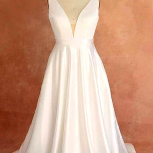 Puede incluir: Vestido de novia blanco sin mangas con escote en V pronunciado y falda fluida. El vestido se exhibe en un maniquí con una etiqueta de talla 20. La tela es lisa y satinada.