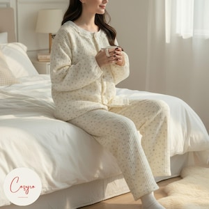 May include: Cream-coloured pyjama set with a floral pattern. The set includes a button-down top and matching trousers. The pyjamas are made of a quilted fabric. The image shows the pyjama set on a bed.
