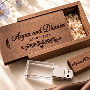 May include: A rectangular wooden sliding box with the engraved text Aryan and Dhania 20 07 2024 and a floral motif. Inside the box is white filler material. A matching wooden USB flash drive with the same engraved text sits beside it.