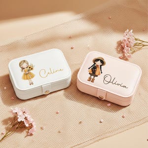 May include: Two rectangular jewelry boxes. One is white with a girl illustration and the name "Celine" in gold script. The other is pink with a girl illustration and the name "Olivia" in black script. Both have a snap closure.