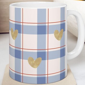 May include: White ceramic mug with a blue and white plaid pattern, red grid lines, and gold heart accents. The mug sits on a beige coaster. A kitchen setting with fruit in a bowl is in the background.