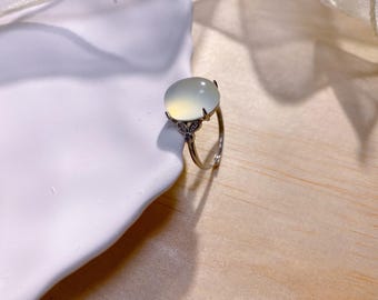 May include: A silver ring with a large, oval, pale green gemstone. The ring's band is thin, and the setting features small, decorative accents. The gemstone has a soft, translucent appearance, and the ring is set against a neutral background.
