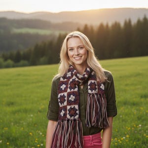 Peut inclure: Une femme porte une écharpe en crochet avec un motif carré dans les tons bordeaux, crème et bleu marine. L'écharpe a de longues franges et est portée sur une chemise verte. L'arrière-plan présente un champ vert et une forêt.