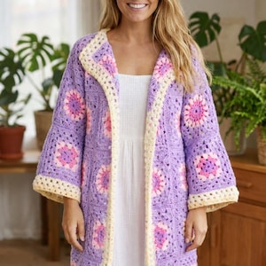 Peut inclure: Longue veste en crochet lilas, ornée de motifs floraux roses et crème. Cette veste ouverte présente des bordures et des manches évasées crème. Le motif en crochet est visible, créant une texture intéressante et un style bohème.