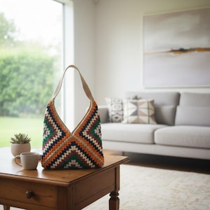Peut inclure: Un sac à bandoulière en crochet fait à la main avec un motif géométrique aux tons orange, vert, marine et crème. Le sac a une sangle beige et est présenté sur une table en bois. Un accessoire élégant.