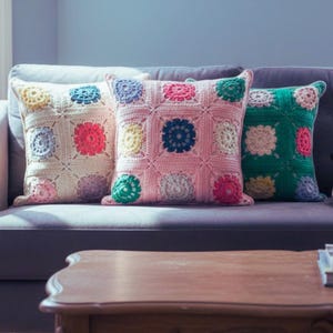 Op de afbeelding: Drie vierkante gehaakte kussens in crème, roze en groen liggen op een grijze bank. Elk kussen heeft een rasterpatroon met kleurrijke bloemmotieven in het midden van elk vierkant. De kussens geven de woonkamer een gezellige, handgemaakte uitstraling.