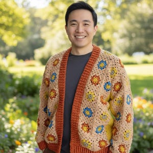 Peut inclure: Un cardigan en crochet beige à motif floral jaune, bleu et orange. Le cardigan a une bordure couleur rouille et est porté sur un t-shirt gris. L'arrière-plan est une scène extérieure floue avec de la verdure.