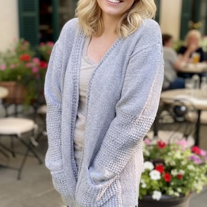 Peut inclure: Un cardigan gris clair avec un col en V et des poches. Le cardigan est en maille et porté sur un haut clair et un jean bleu. Les manches ont un motif texturé.