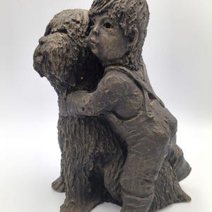 Puede incluir: Una escultura texturizada de color marr&oacute;n oscuro que representa a un ni&ntilde;o con peto abrazando a un perro de pelo peludo. La figura muestra al ni&ntilde;o con los brazos alrededor del cuello del perro, capturando una interacci&oacute;n cercana.