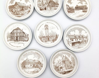 May include: A collection of nine white ceramic plates, each featuring a sepia-toned illustration of a building or landmark. The plates have a circular shape and are arranged in a grid pattern. The illustrations include text.