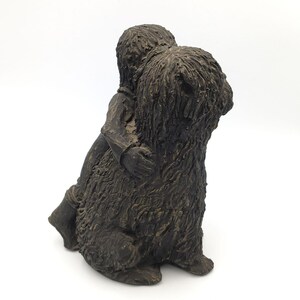 Puede incluir: Una escultura texturizada de color marr&oacute;n oscuro que representa a una figura abrazando a un perro de pelo largo y desgre&ntilde;ado. La pieza presenta un acabado monocrom&aacute;tico con surcos profundos y detalles en relieve que definen el pelaje.