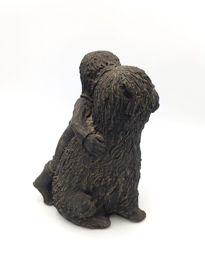 Puede incluir: Una escultura texturizada de color marr&oacute;n oscuro que representa a una figura abrazando a un perro de pelo largo y desgre&ntilde;ado. La pieza presenta un acabado monocrom&aacute;tico con surcos profundos y detalles en relieve que definen el pelaje.