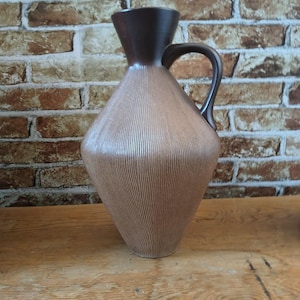 Può includere: Una brocca in ceramica marrone con un motivo a linee verticali e testurizzate. La brocca ha un manico marrone scuro e una parte superiore svasata marrone scuro. La brocca è appoggiata su una superficie di legno contro un muro di mattoni.
