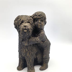 Puede incluir: Una escultura en tonos bronce que representa a un ni&ntilde;o con el pelo corto y peto, abrazando a un perro grande de pelaje desgre&ntilde;ado. La figura presenta un acabado monocrom&aacute;tico y texturizado con reflejos marr&oacute;n oscuro y tonos bronce.