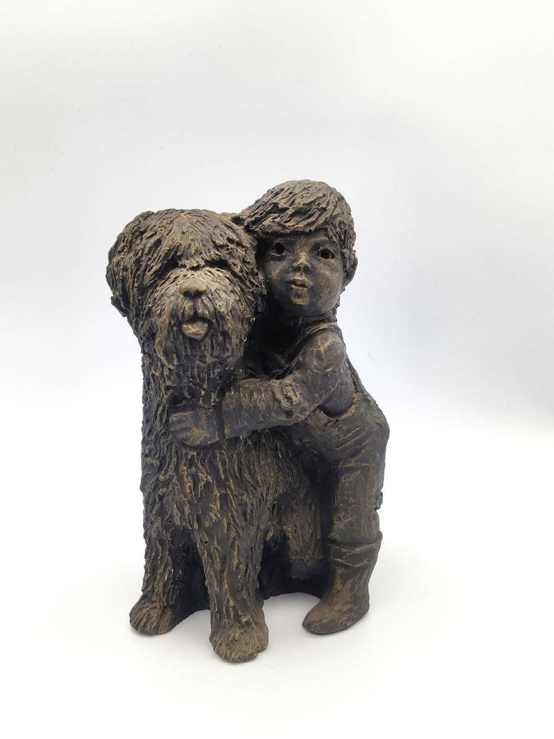 Puede incluir: Una escultura en tonos bronce que representa a un ni&ntilde;o con el pelo corto y peto, abrazando a un perro grande de pelaje desgre&ntilde;ado. La figura presenta un acabado monocrom&aacute;tico y texturizado con reflejos marr&oacute;n oscuro y tonos bronce.