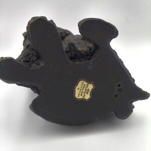 Puede incluir: Una base de figura de color marr&oacute;n oscuro y textura rugosa con una forma org&aacute;nica irregular. En el centro hay una peque&ntilde;a pegatina en forma de escudo con el texto Gilde Handwerk en letras negras sobre un fondo dorado y un icono de corona.