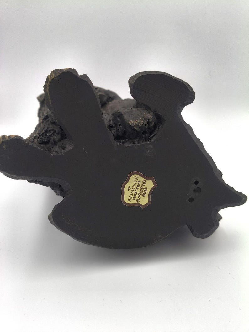 Puede incluir: Una base de figura de color marr&oacute;n oscuro y textura rugosa con una forma org&aacute;nica irregular. En el centro hay una peque&ntilde;a pegatina en forma de escudo con el texto Gilde Handwerk en letras negras sobre un fondo dorado y un icono de corona.