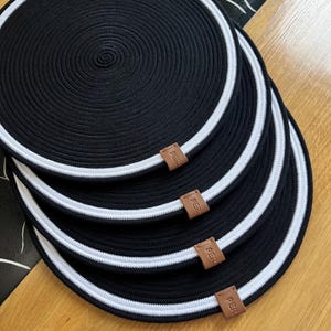 Puede incluir: Un conjunto de cuatro manteles individuales circulares negros con un borde blanco. Cada mantel individual tiene una etiqueta de cuero marrón con la palabra "PER". Los manteles individuales están hechos de un material tejido y están colocados sobre una superficie de madera.