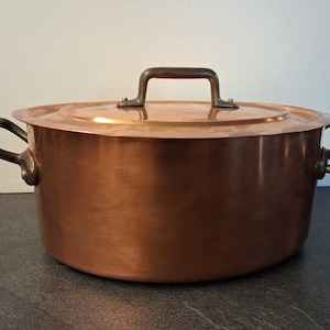 May include: A large copper pot with a matching lid. The pot has two side handles and a handle on the lid. The copper has a warm, reddish-brown color. The handles are a darker, contrasting color.