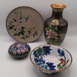 May include: A collection of decorative cloisonné pieces, including a round plate with pink floral and bird designs, a vase with a floral motif, a small round box, and a bowl with bird and floral patterns. The pieces feature gold trim and intricate detailing.