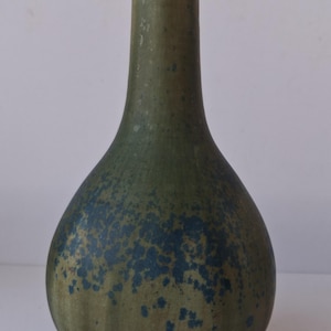Op de afbeelding: Een groene keramische vaas met een gespikkeld blauw patroon. De vaas heeft een afgeronde basis die taps toeloopt naar een smalle hals. De kleur is een gedempt groen met donkerdere blauwe spikkels, waardoor een textuur ontstaat. De vaas is een decoratief item.