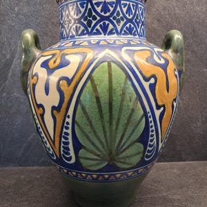 May include: A decorative ceramic vase with two handles. The vase features a green, blue, and yellow floral design. The top and base are green, with a blue and white pattern around the neck. The body has a large green leaf design.