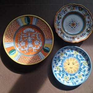 Può includere: Tre piatti decorativi in ceramica con disegni colorati. Uno presenta uno sfondo arancione con un motivo floreale, un altro ha un disegno di sole blu e giallo e il terzo ha un motivo floreale con un bordo smerlato.