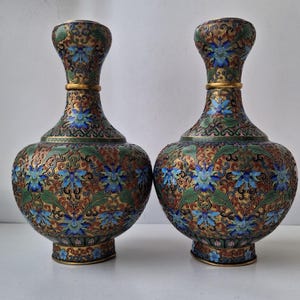 Pode incluir: Dois vasos ornamentados com corpo bulboso e pescoço esguio. Os vasos apresentam um padrão floral detalhado em azul, verde, vermelho e dourado. Uma faixa dourada acentua o pescoço de cada vaso. Os vasos são provavelmente itens decorativos.