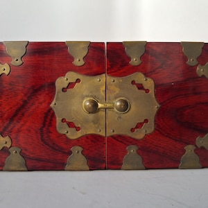 May include: A rectangular, wooden box with a rich, reddish-brown finish. The box is adorned with ornate, brass-colored metal corner brackets and a central clasp. The wood grain is visible, adding texture to the design. The box is closed.