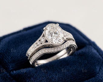 May include: A silver-toned engagement ring set featuring an oval-cut center stone held by four prongs. The band displays intricate milgrain detailing, engraved patterns, and small round accent stones along the shank and the matching curved band.