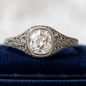 May include: A silver-toned ring featuring a central cushion-cut clear stone set within a decorative filigree band. The metalwork displays intricate scroll patterns and milgrain detailing along the edges, creating a textured appearance.