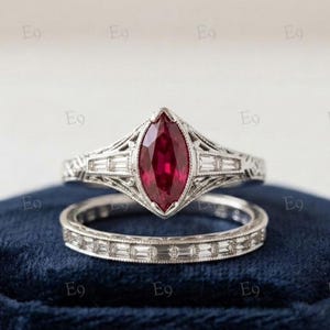 Puede incluir: Un conjunto de anillos en tono plateado con un rubí rojo intenso en talla marquesa como piedra central, rodeado de pequeñas piedras claras en talla baguette. La banda presenta un intrincado trabajo de filigrana y una alianza a juego.
