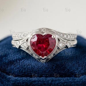 Puede incluir: Un anillo de tono plateado con una prominente piedra preciosa roja en forma de corazón en el centro. La banda está detallada con pequeñas piedras transparentes incrustadas a lo largo del marco metálico, que presenta una textura milgrain grabada.