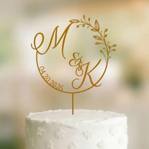 May include: A gold-coloured cake topper featuring a circular frame with a leaf design, the letters M and K, an ampersand, and the date 04.20.2026. The topper is mounted on a single thin stem and is positioned atop a white two-tiered frosted cake.