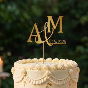 May include: A gold-colored cake topper featuring the letters A and M connected by an ampersand symbol, with the date 5.15.2026 displayed underneath. The topper is positioned on top of a two-tiered white cake decorated with piped frosting and gold leaf.