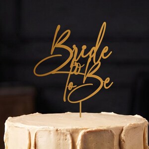 Könnte beinhalten: Ein goldfarbener Tortenstecker mit dem geschriebenen Text Bride to Be, der auf einem dünnen vertikalen Stiel befestigt ist. Der Tortenstecker steht aufrecht in der Mitte einer runden Torte mit einer strukturierten, hellbeigen Glasur.